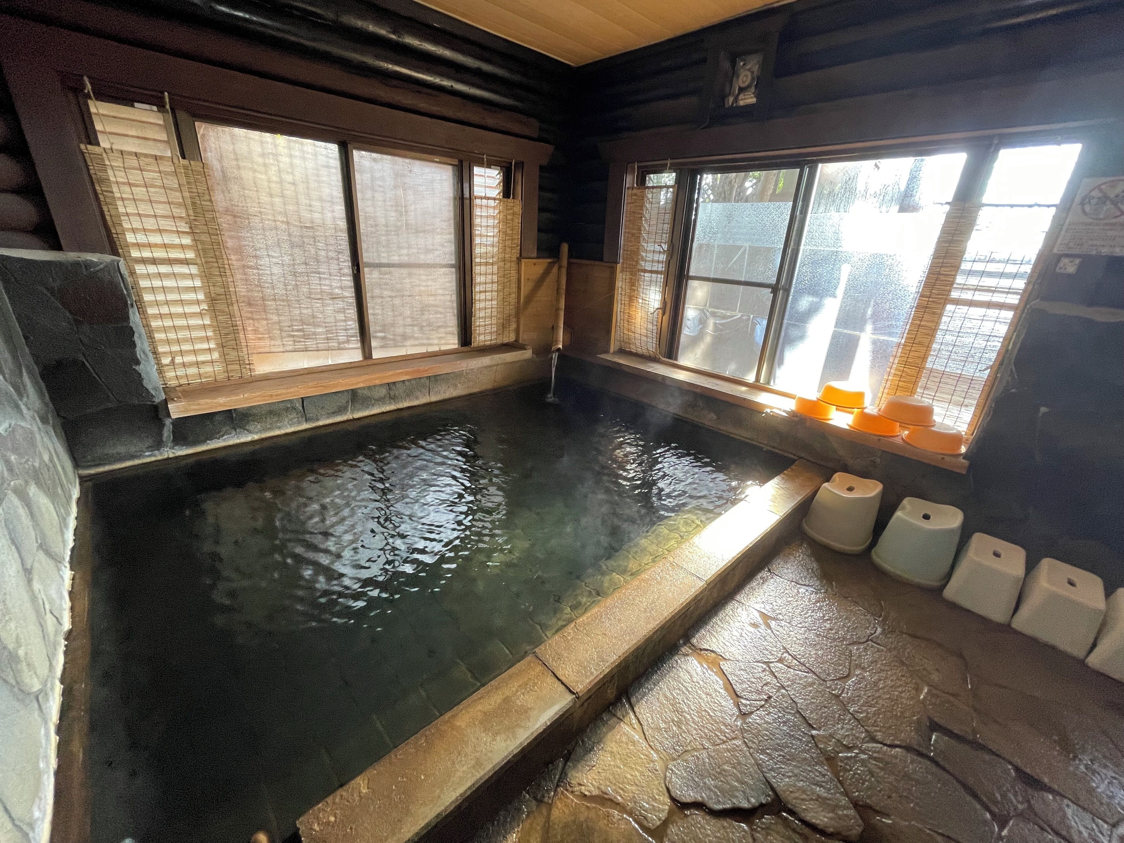 Un onsen japonais traditionnel, avec un bain en pierre, des portes coulissantes en bois et une vue sur l’extérieur par les fenêtres.