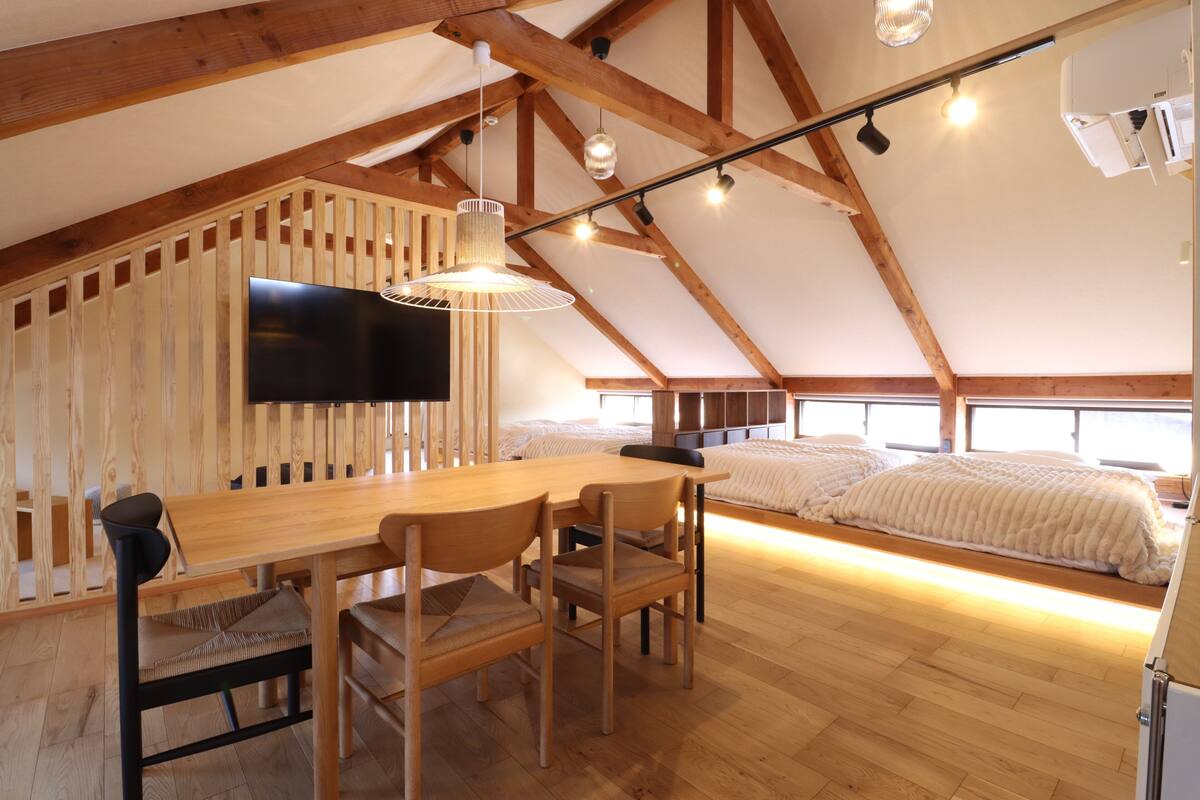 A spacious loft with a high ceiling, wooden beams, a dining area with a wooden table and chairs, a large flat-screen TV, and a bed with a white duvet.
