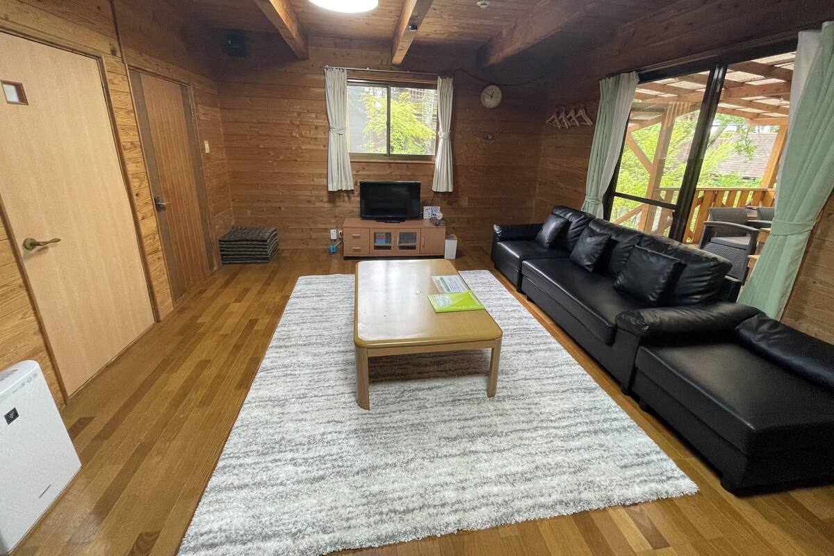 A cozy cabin interior with a wooden floor, a black leather sofa, a wooden coffee table, a TV on a wooden stand, and a large window with curtains.