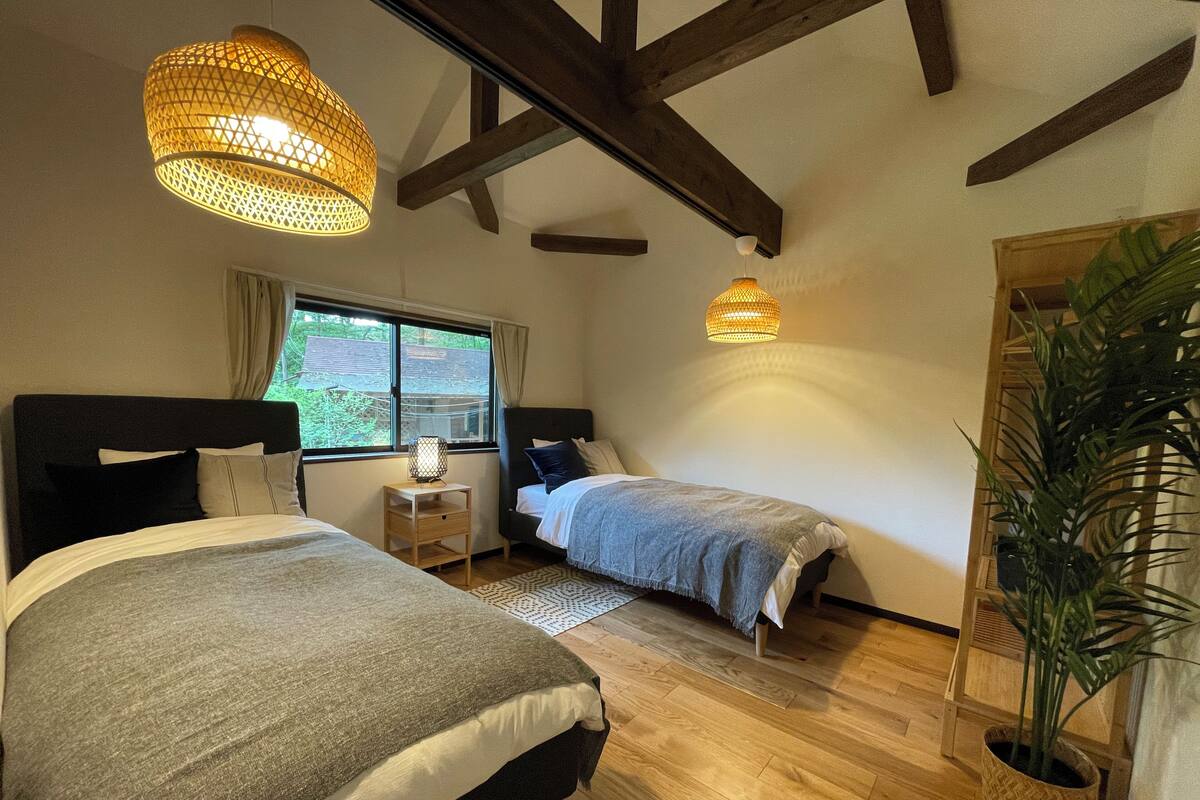 A bedroom with two beds, wooden beams, a window with curtains, a bedside table with a lamp, and a potted plant.