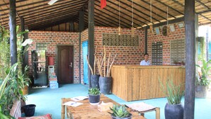 Un espacio al aire libre techado con pared de ladrillo, mesas de madera, plantas en macetas y una zona de bar.