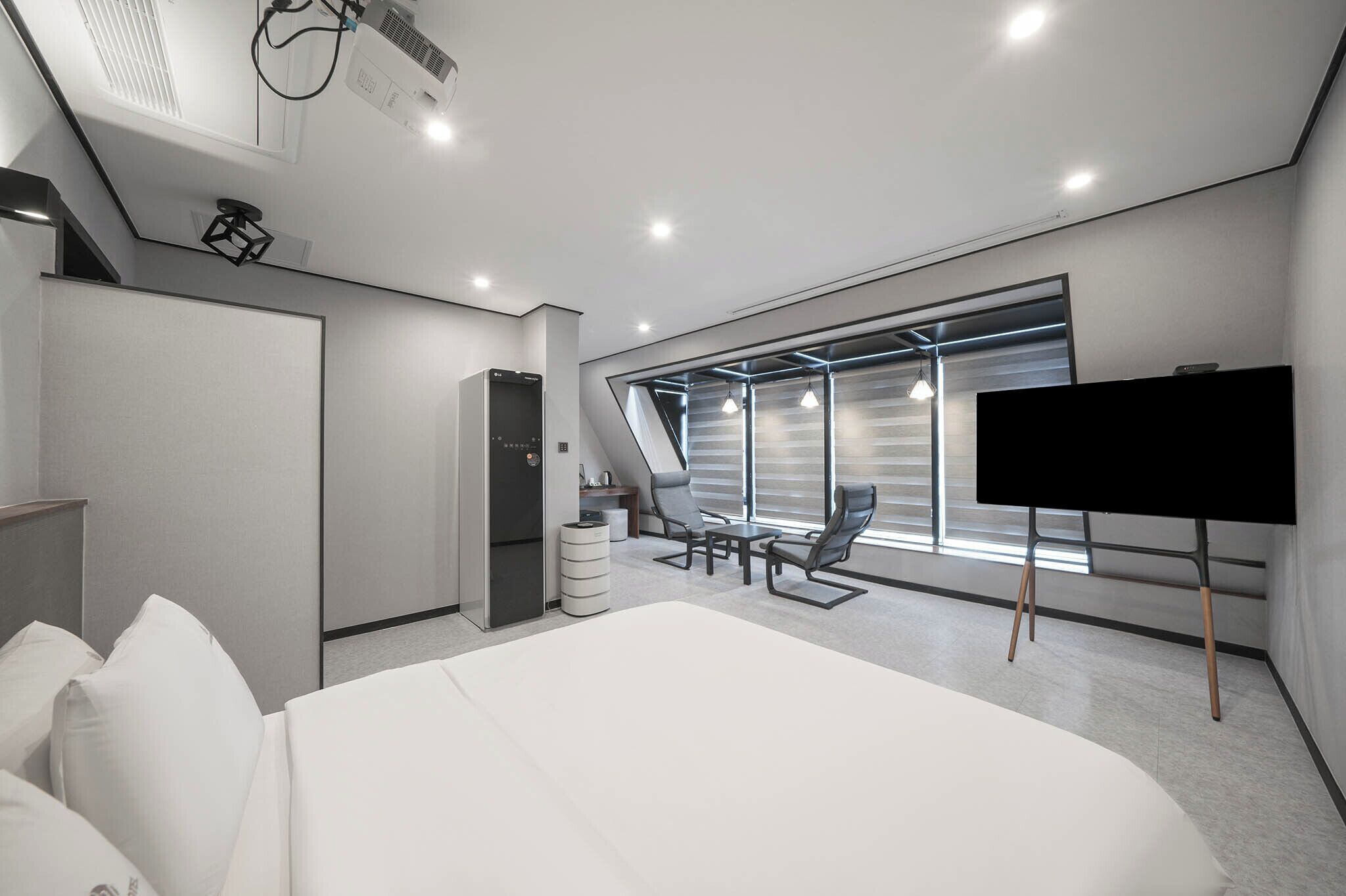 A modern hotel room with a large bed, a desk, a chair, and a TV. There is a window with blinds and a view of the outside.