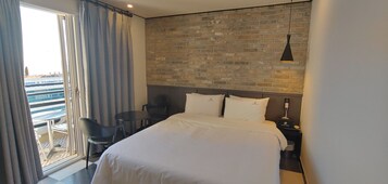 Habitación de hotel con una cama grande, un escritorio y una silla. Se observa una pared de ladrillo detrás de la cama y una ventana con cortinas.