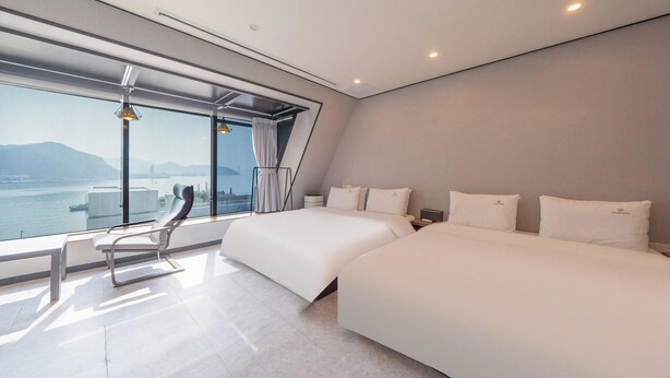 A modern hotel room with a large window offering a city view, two beds, a chair, and a nightstand. - Lucas Hotel (Changwon)