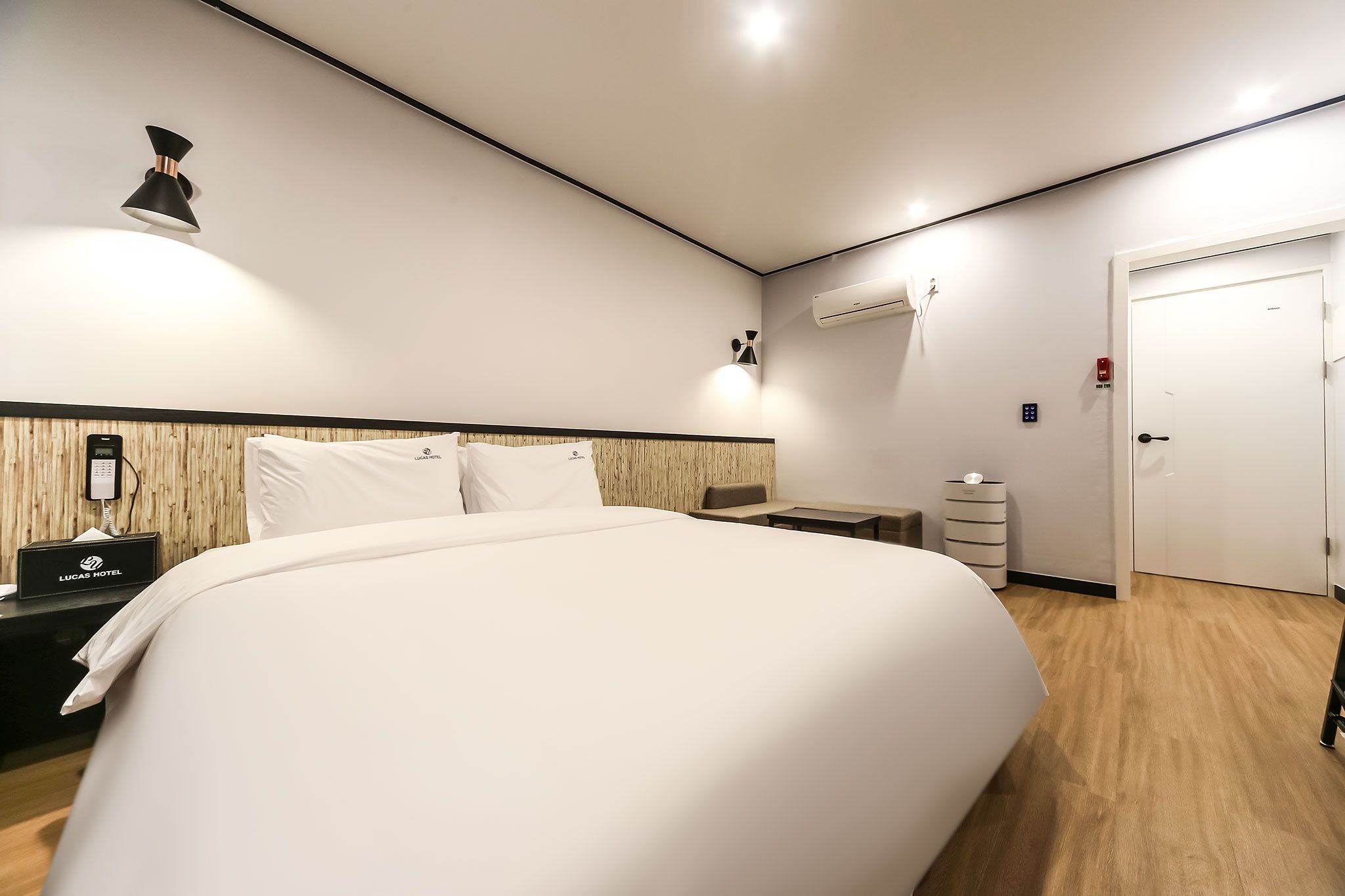 A hotel room with a large bed, a bedside table with a phone, a wall-mounted light, a small desk, a chair, a wardrobe, and a door.