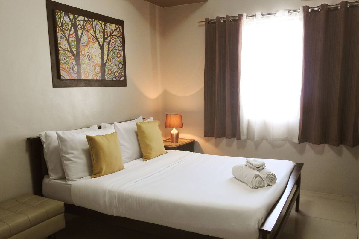 A neatly made bed with white and yellow pillows, a bedside lamp, and a framed artwork on the wall.