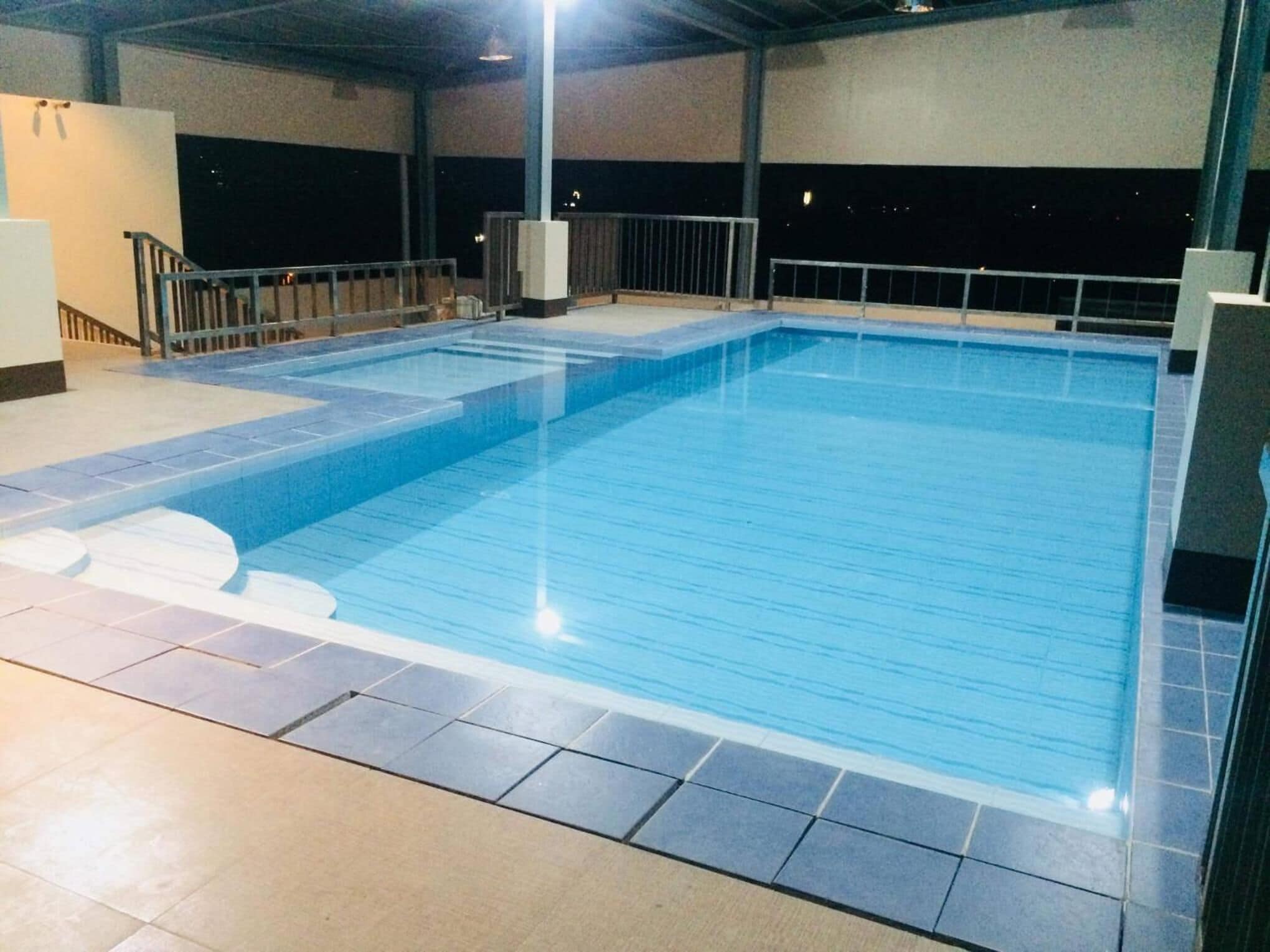 An indoor swimming pool with clear blue water, tiled flooring, and a metal railing around the perimeter.