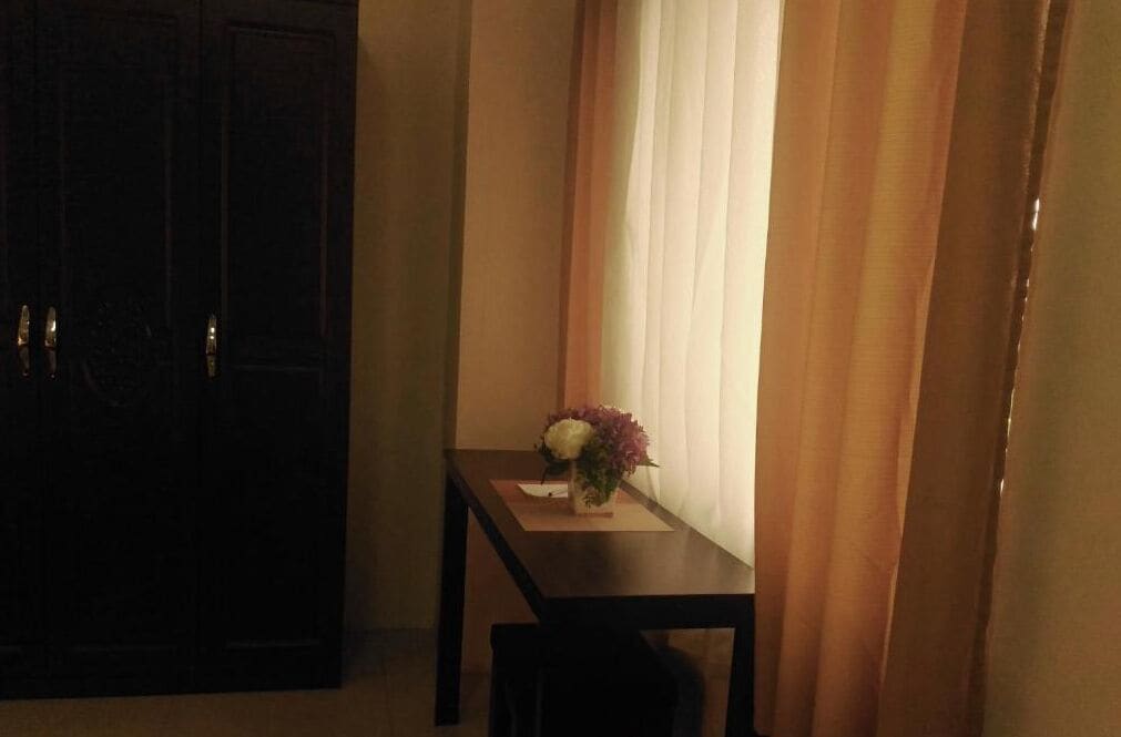 Une pièce avec une armoire en bois sombre, une petite table sur laquelle est posé un vase de fleurs et des rideaux.