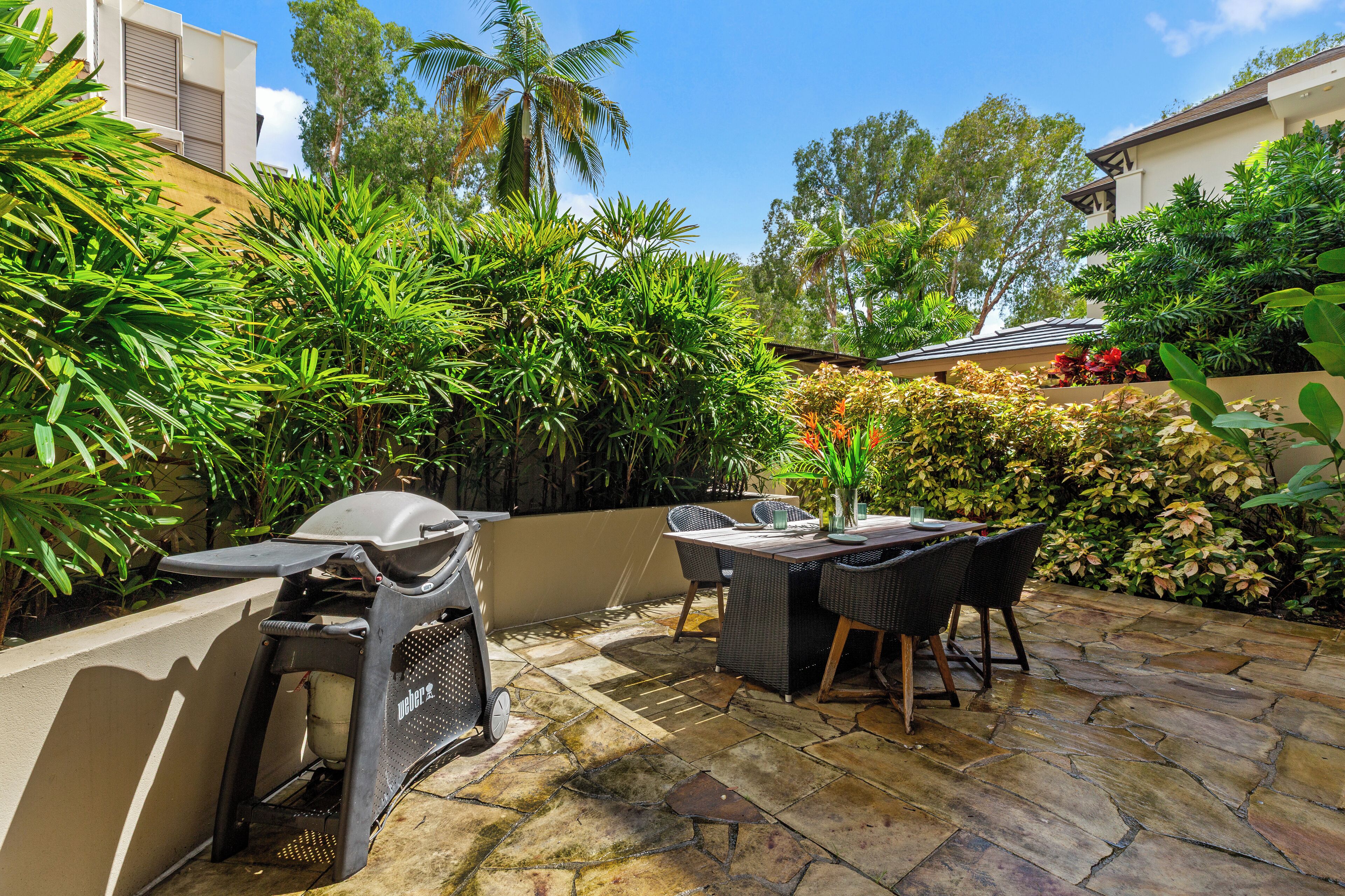 An outdoor patio with a grill, dining table, and chairs, surrounded by lush greenery and a stone floor.