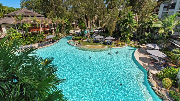 A large, clear swimming pool surrounded by lush greenery and palm trees, with lounge areas and a small island.