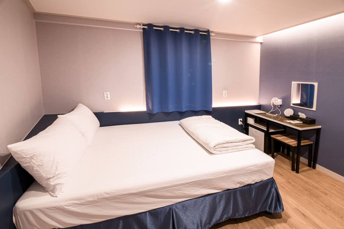A neatly made bed with a blue curtain, a desk with a mirror, and a chair.