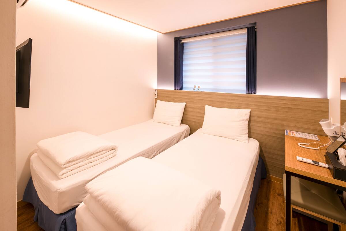 A hotel room with two single beds, a wooden headboard, a bedside table with a lamp, a window with blinds, and a wall-mounted TV.