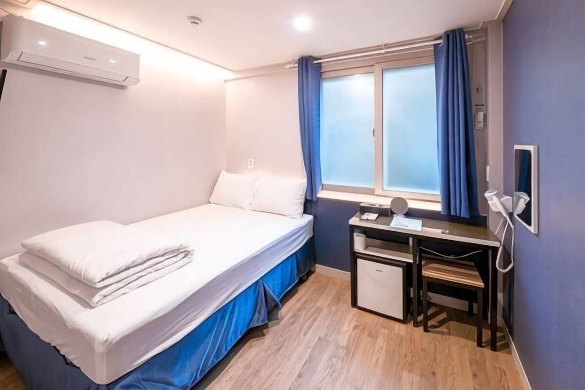A single bed with a white mattress and pillows, a desk with a chair, a small cabinet, and a window with blue curtains.