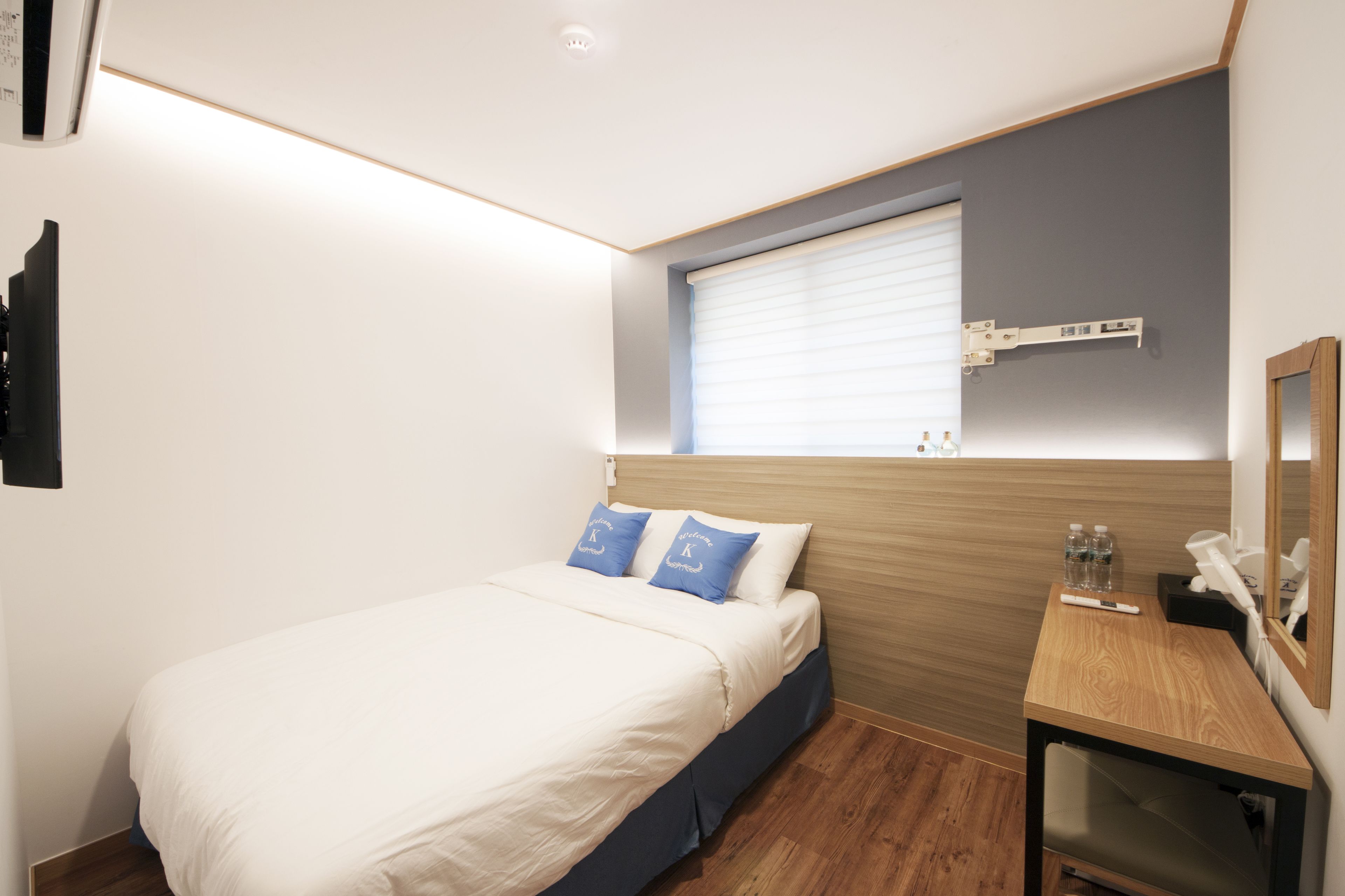 A bedroom with a bed, a desk, a window with blinds, and a mounted shelf with bottles.