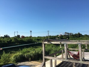 A wooden deck with a hammock and a rope barrier, overlooking a lush green landscape with a clear blue sky. - OKINAWA MOTOBU GUESTHOUSE - Hostel (Motobu)