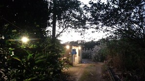 A narrow pathway leading to a small, illuminated structure with a hanging item, surrounded by dense foliage. - OKINAWA MOTOBU GUESTHOUSE - Hostel (Motobu)