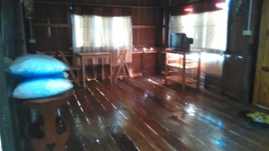 Une pièce avec un plancher en bois, une table entourée de chaises, un téléviseur posé sur un meuble et une fenêtre avec des rideaux.