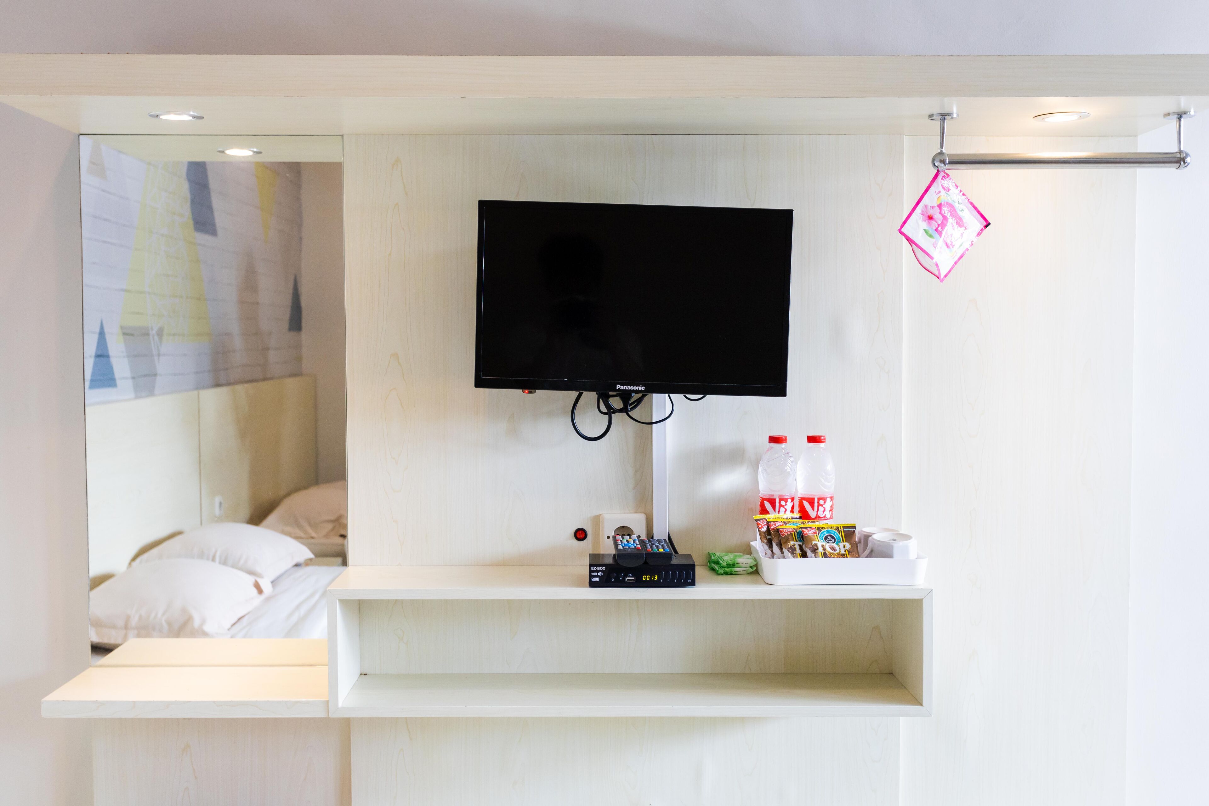 A hotel room with a mounted television, a bedside table with a bottle of soda, and a small shelf with various items.
