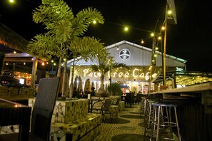 An outdoor seating area with stone planters and metal chairs, a thatched-roof structure, and a building with a sign reading "Palm Tree Cafe". - Batuque Town Villa (Batu)
