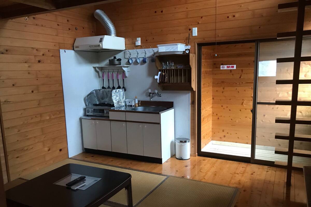 Una cocina compacta con estufa, lavamanos y armarios de madera. Hay una mesa pequeña con un control remoto y un bote de basura.