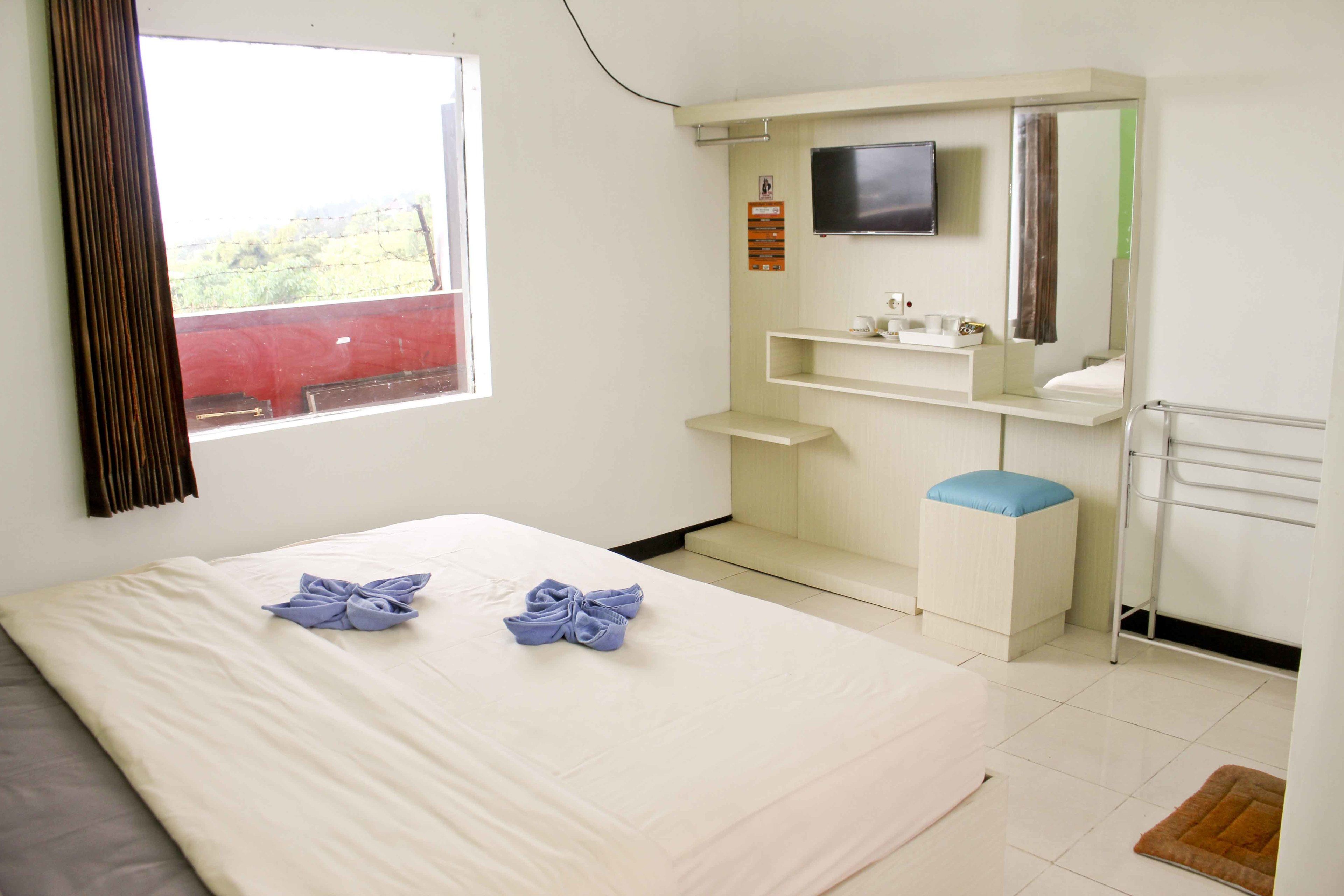 A hotel room with a bed, a TV mounted on the wall, a small shelf with toiletries, and a window with a curtain.
