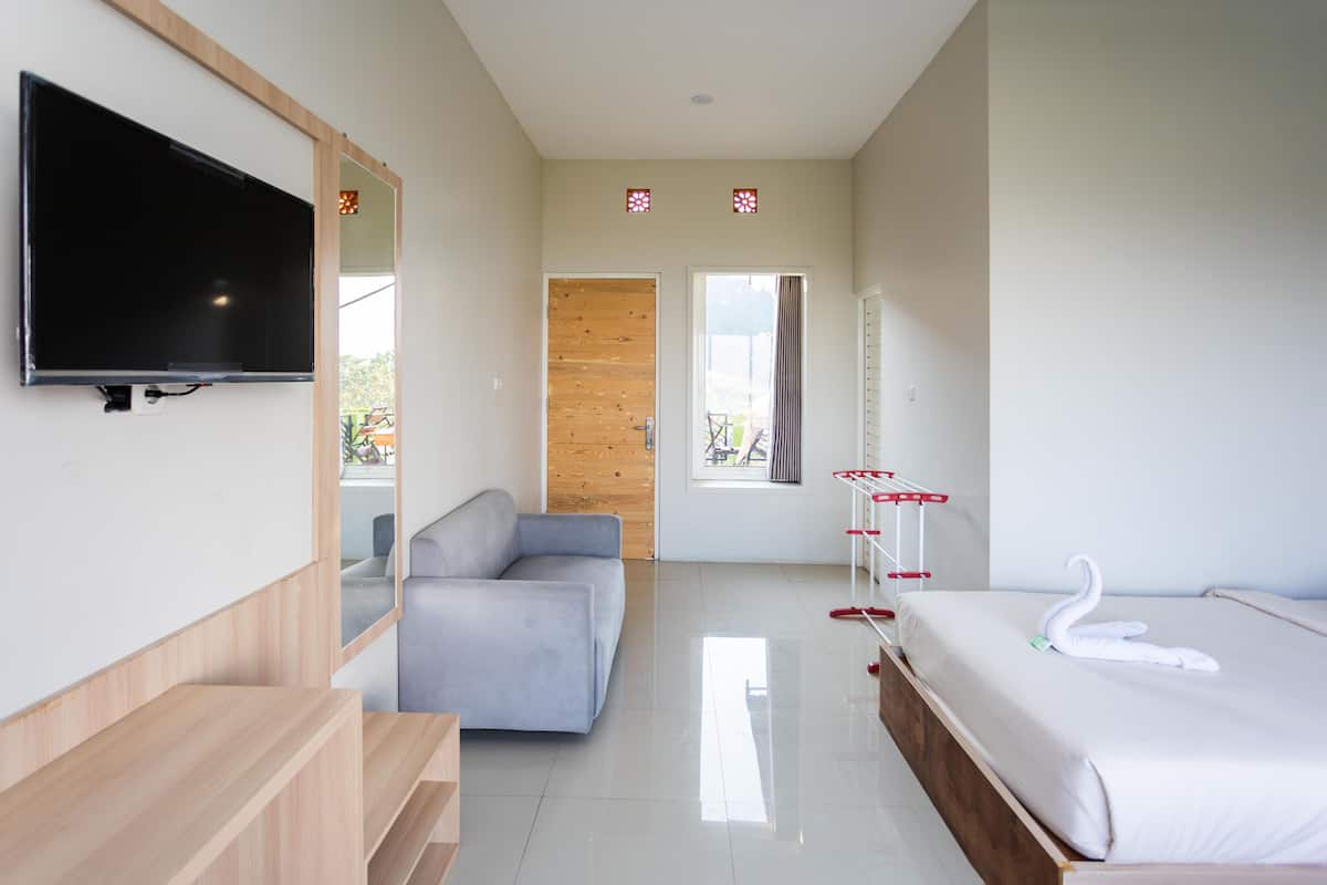 Una habitación de hotel moderna con una televisión de pantalla plana, un sofá gris, una puerta de madera, una ventana con cortinas, una cama con sábanas blancas y una decoración con forma de cisne, y un espejo en la pared.