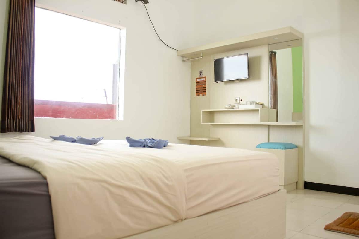 Habitación de hotel con cama, televisor montado en la pared, estante pequeño con artículos de aseo y ventana con cortinas.
