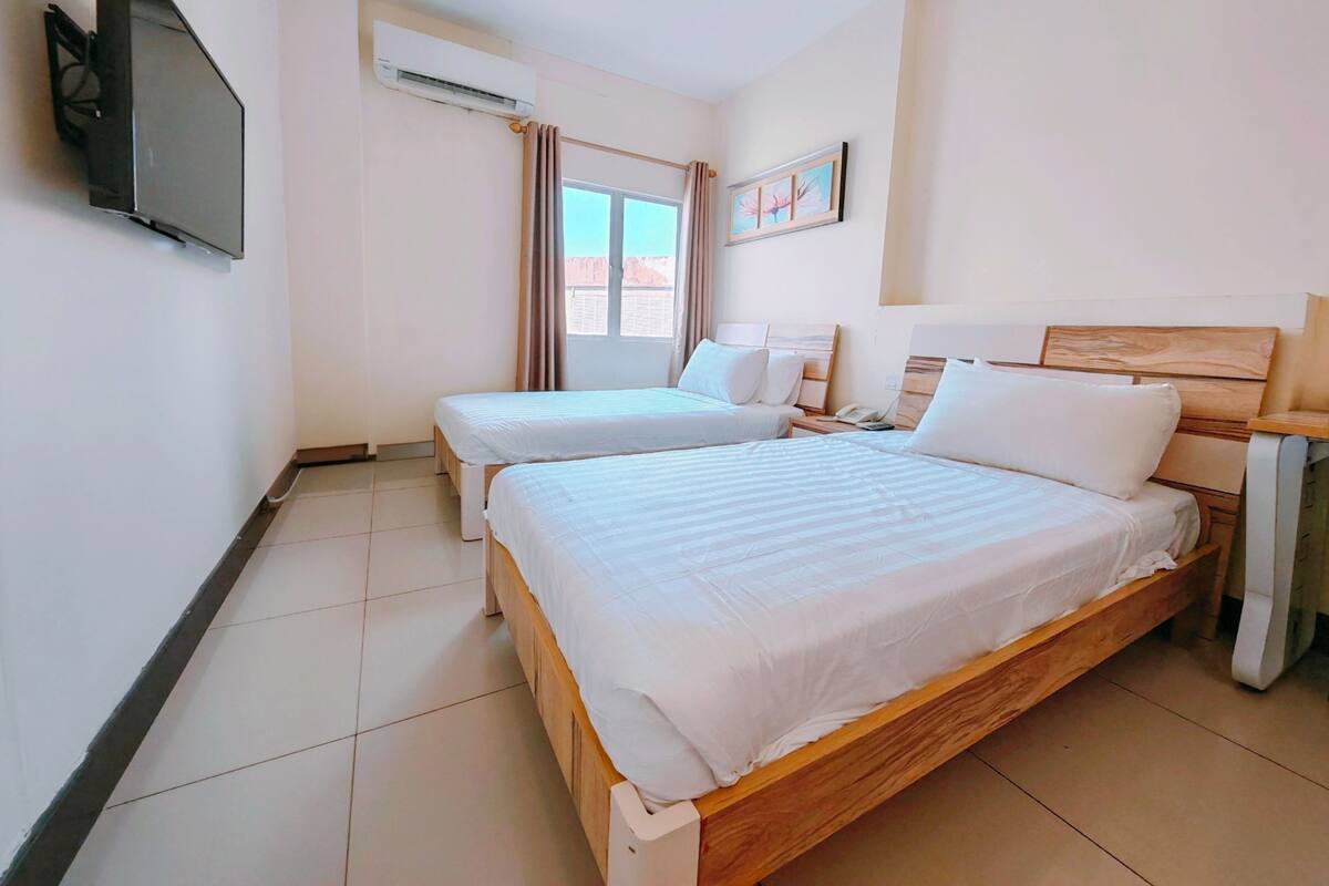A hotel room with two beds, a TV, an air conditioner, a window with curtains, and a painting on the wall.