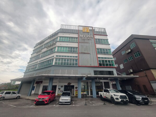 Un hotel de varios pisos con fachada moderna, unidades de aire acondicionado y un estacionamiento con varios autos.