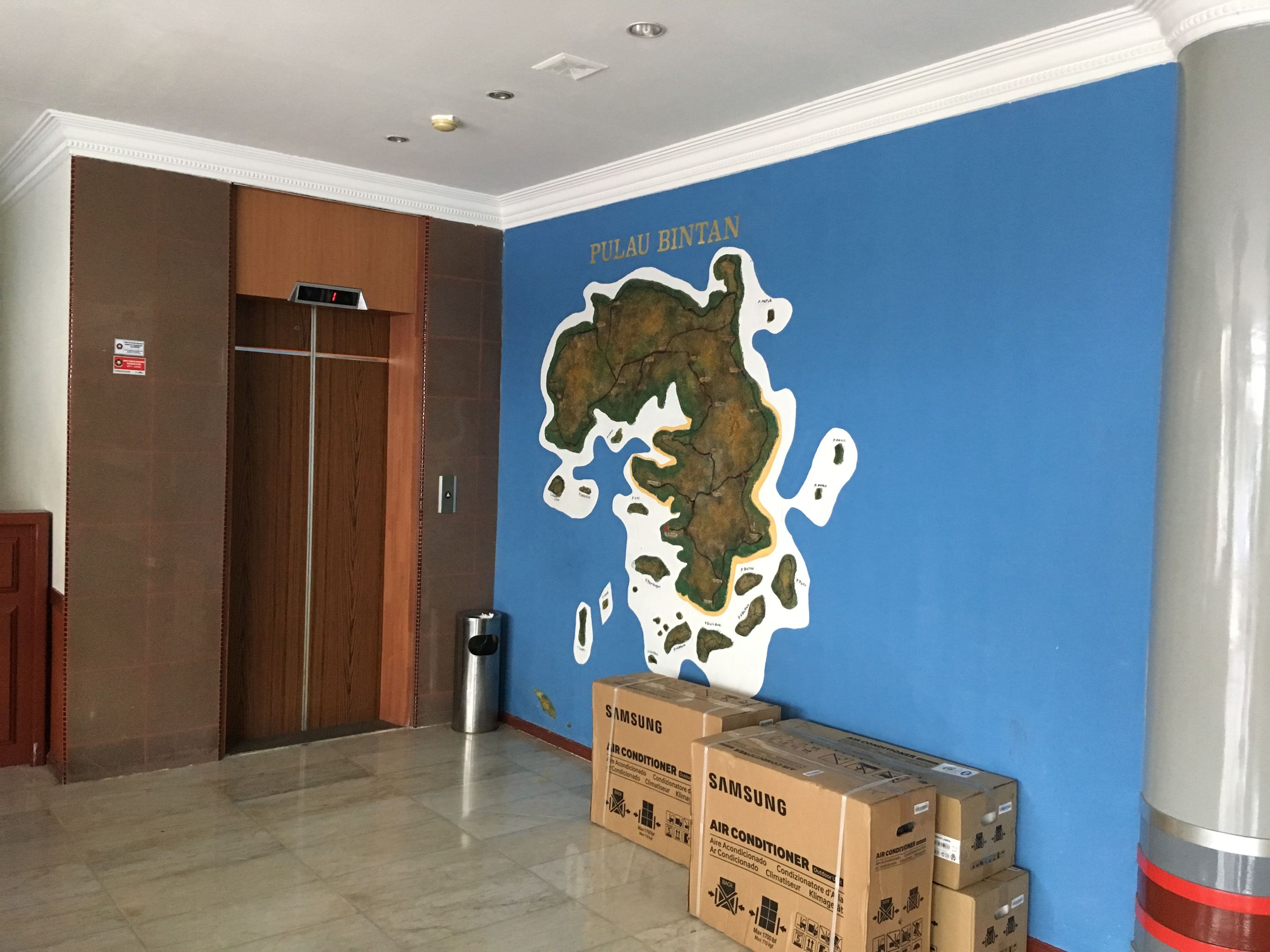 Una habitación con una pared azul que muestra un mapa de Pulau Rintan, una puerta de madera y cajas de aire acondicionado Samsung en el suelo.
