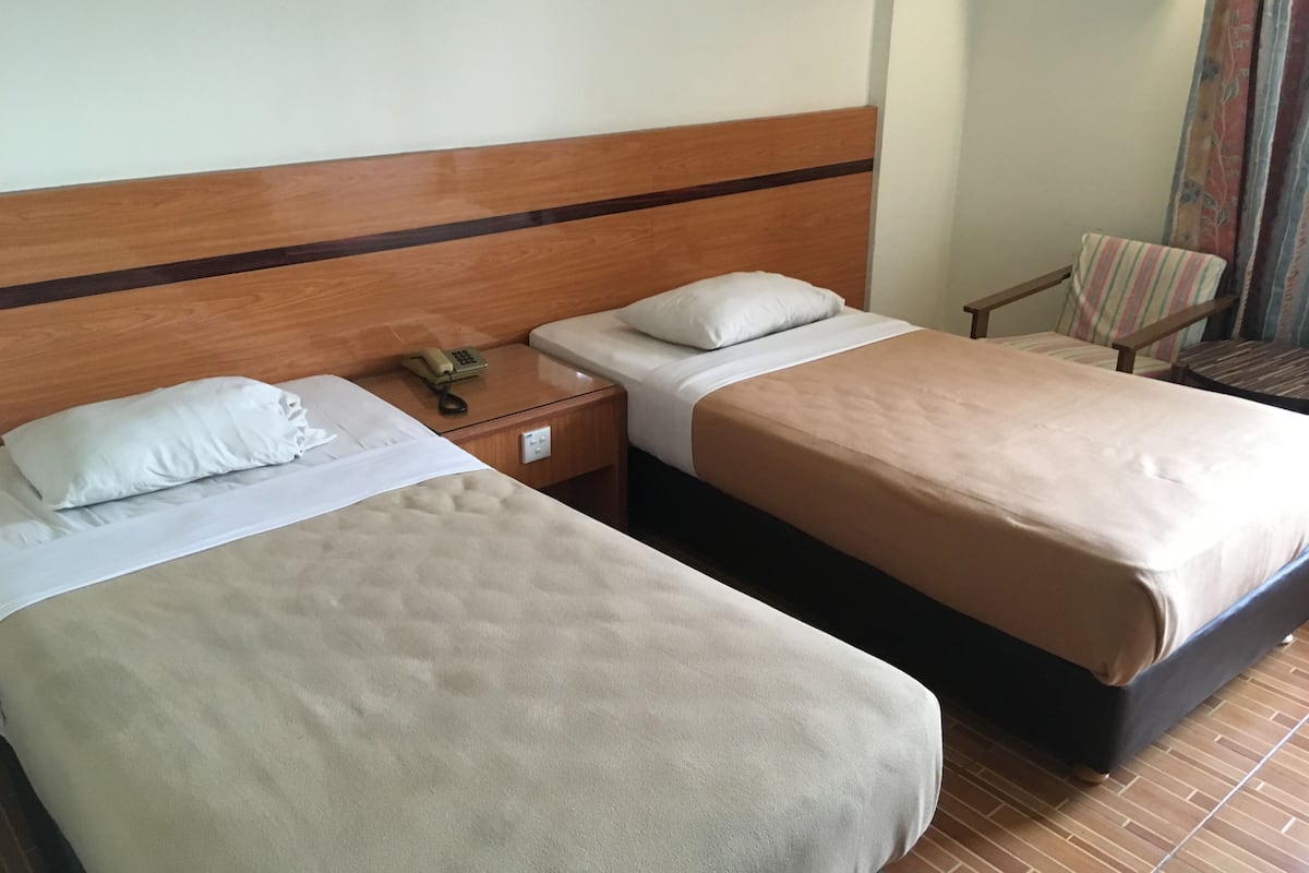A hotel room with two single beds, a wooden headboard, a bedside table with a phone, a chair, and a window with curtains.