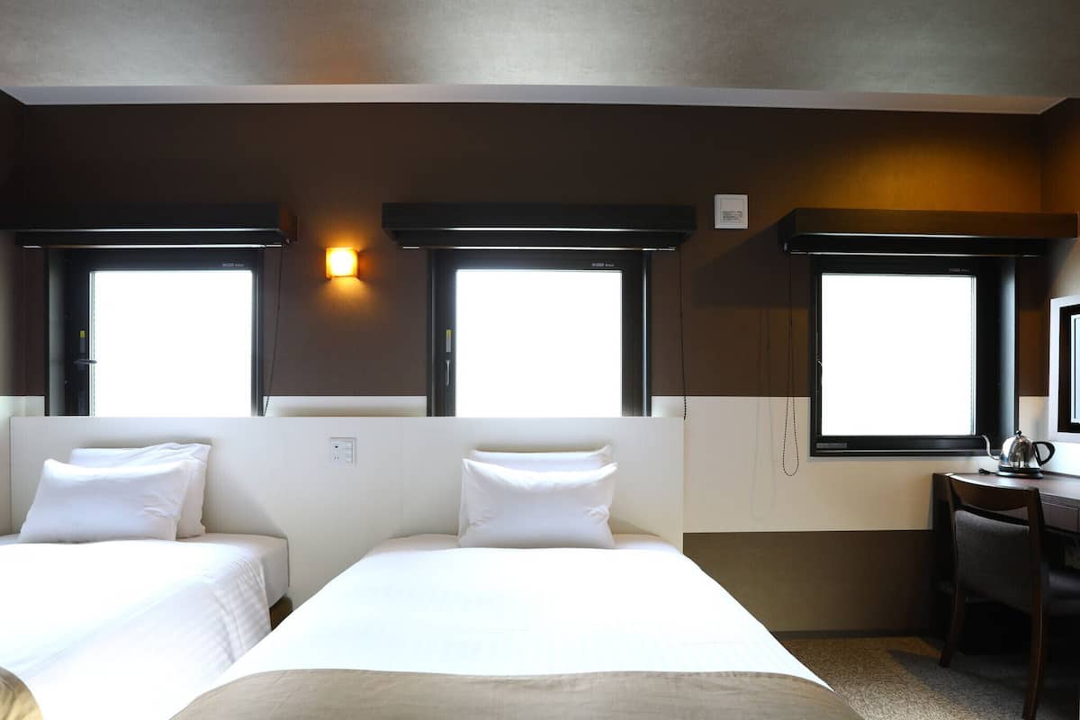Une chambre d’hôtel avec deux lits, un bureau et une chaise. Elle dispose de trois fenêtres qui laissent entrer la lumière naturelle.