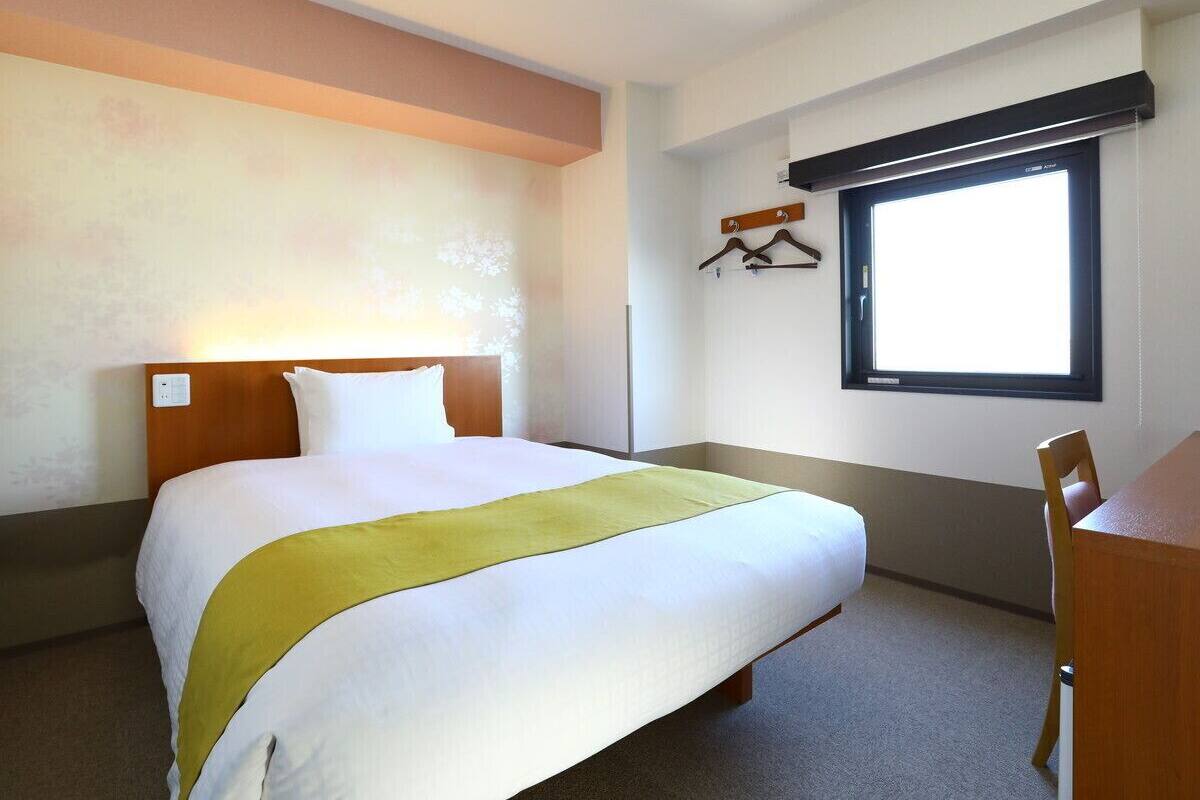 Une chambre d’hôtel avec un lit, un bureau, une chaise, une fenêtre et un porte-manteau.