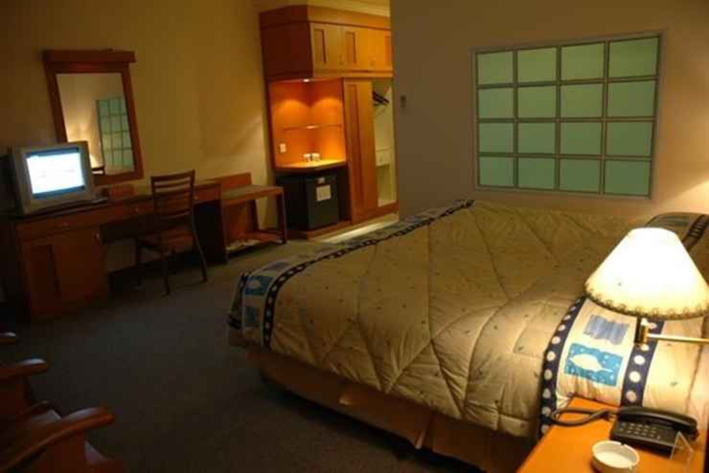 A hotel room with a bed, a desk with a computer, a chair, a lamp, and a small table with a phone and a cup.