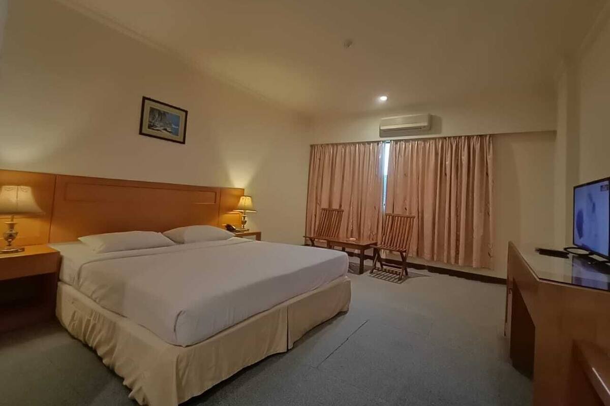 A hotel room with a large bed, two bedside tables with lamps, a desk with a computer, and a window with curtains.