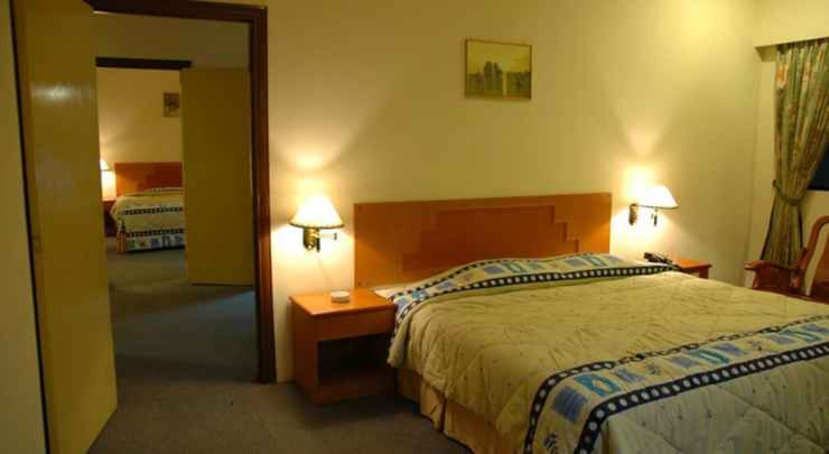 Habitación de hotel con cama, lámparas de noche, mesita de noche y una puerta que da a otra habitación.