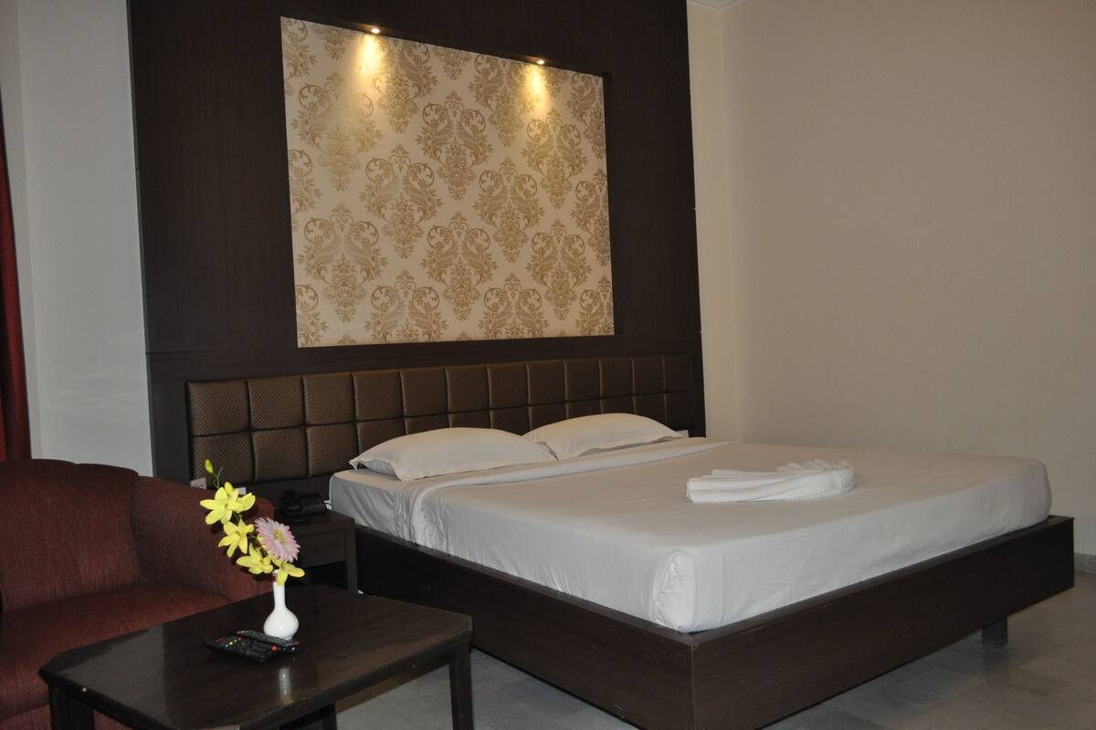 Quarto de hotel com uma cama grande, cabeceira escura, mesa de cabeceira, controle remoto e um vaso com flores.