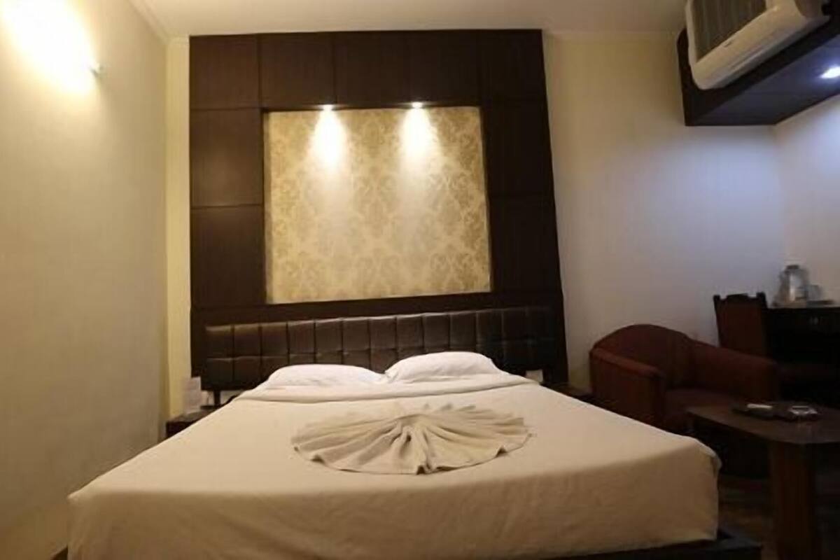 Quarto de hotel com cama, cabeceira iluminada, janela com cortina estampada e escrivaninha com cadeira.