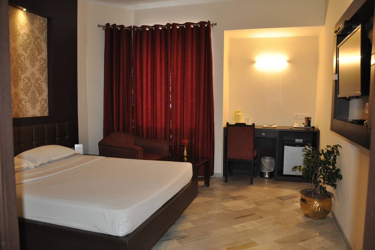 Quarto de hotel com cama, escrivaninha, cadeira, lixeira pequena, planta em vaso e espelho na parede.