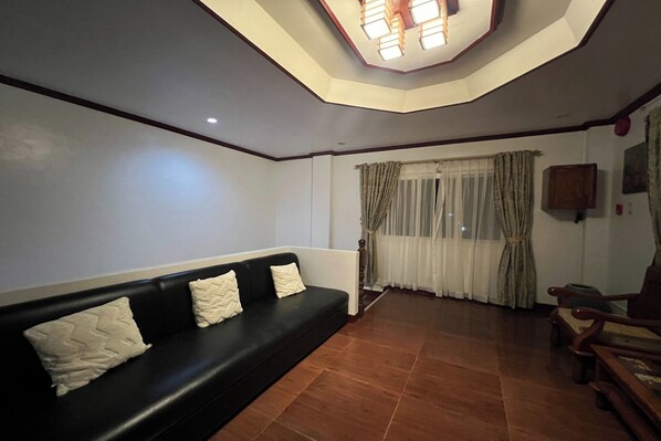 A living room with a black sofa, wooden furniture, a ceiling light fixture, and a window with curtains.