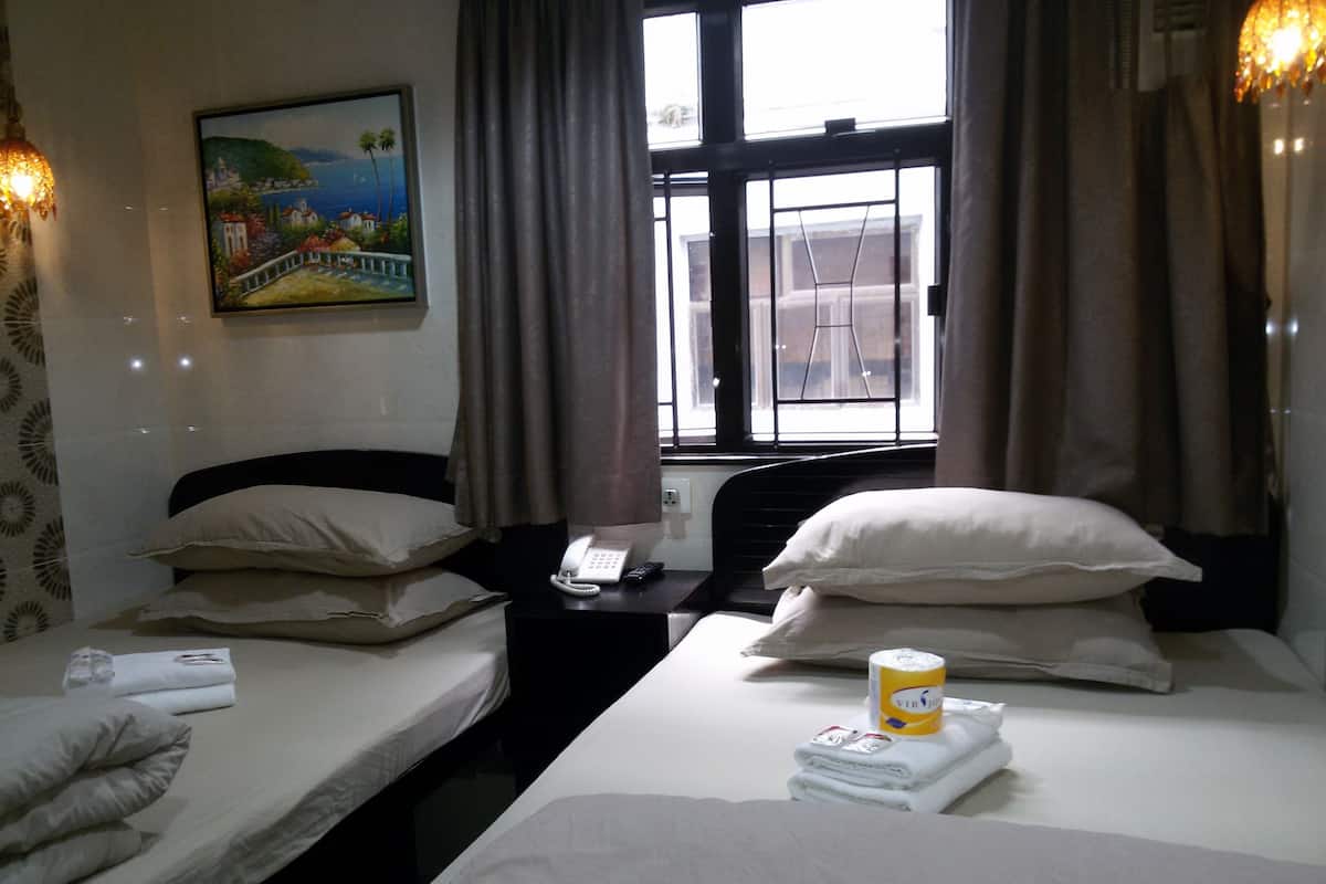 A hotel room with two beds, a painting on the wall, a window with curtains, and a bedside table with a phone and a cup.