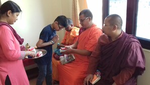 Un gruppo di persone in una stanza con una finestra, una statua di Buddha e un vassoio con degli oggetti.