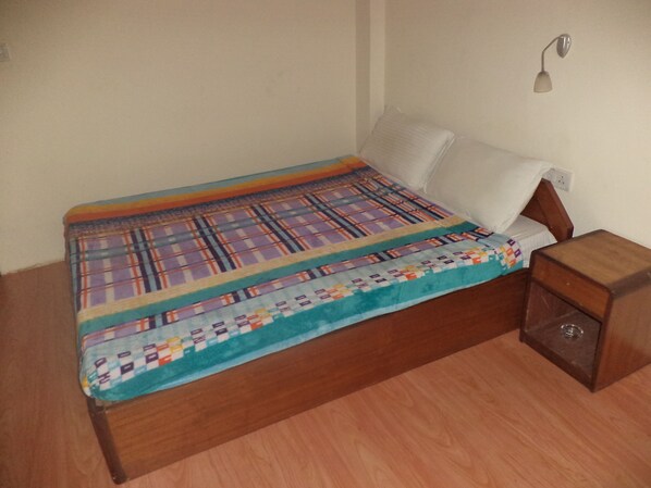 A small, neatly made bed with a colorful quilt, a wooden nightstand with a lamp, and a plain wall.