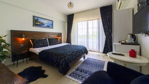 A bedroom with a wooden headboard, a bed with patterned bedding, a small bedside table with a remote control, a TV, and a window with sheer curtains.