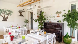 Zona pranzo con tavoli e sedie bianche, un pianoforte e piante ornamentali.