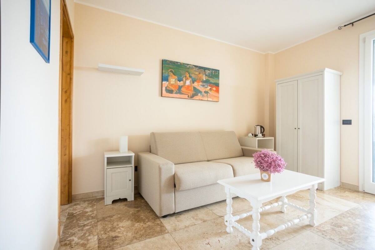 A living room with a beige sofa, a white coffee table with a vase of flowers, a painting on the wall, and a white cabinet.