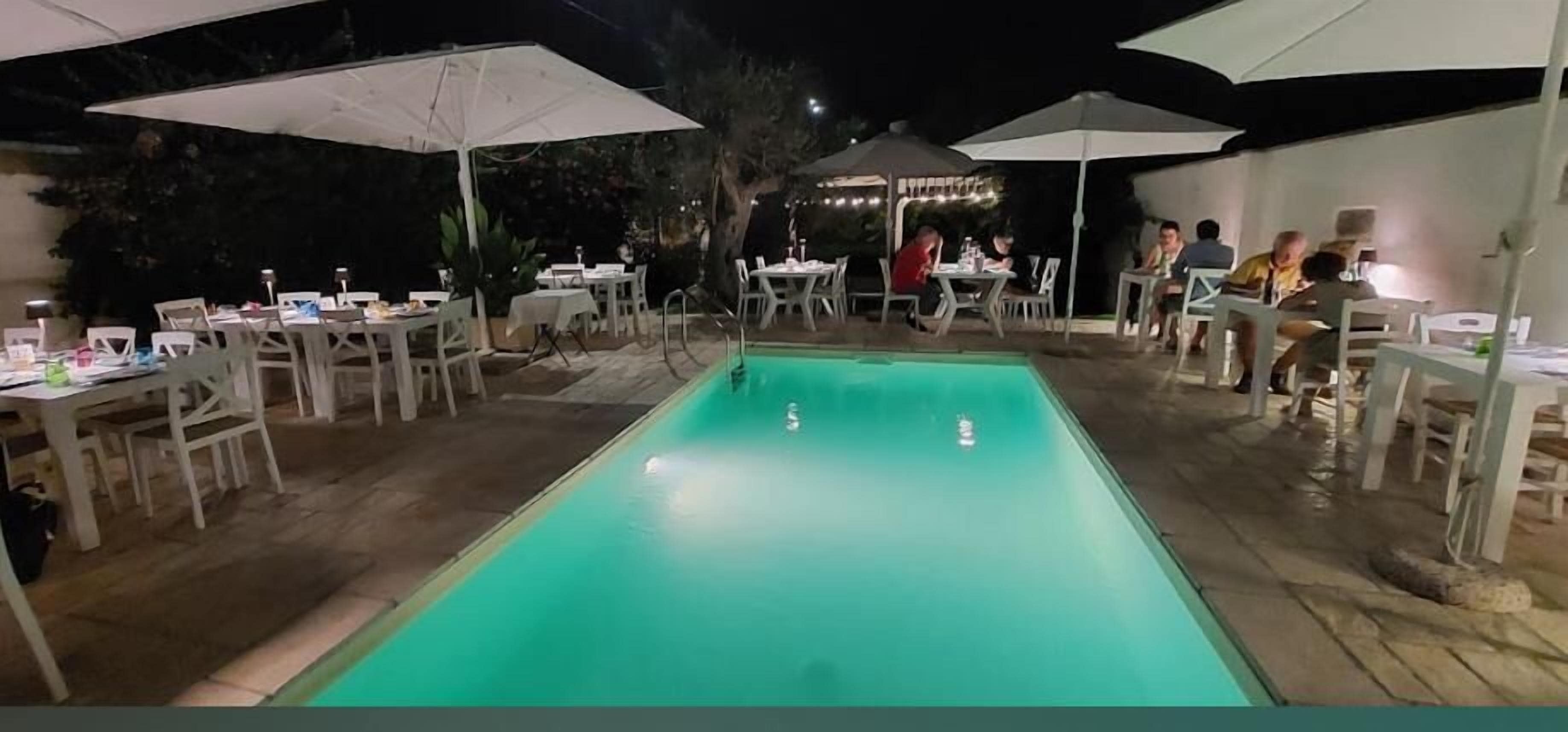 Un espace extérieur avec une piscine, des tables et des chaises sous des parasols, le tout entouré d’arbres.