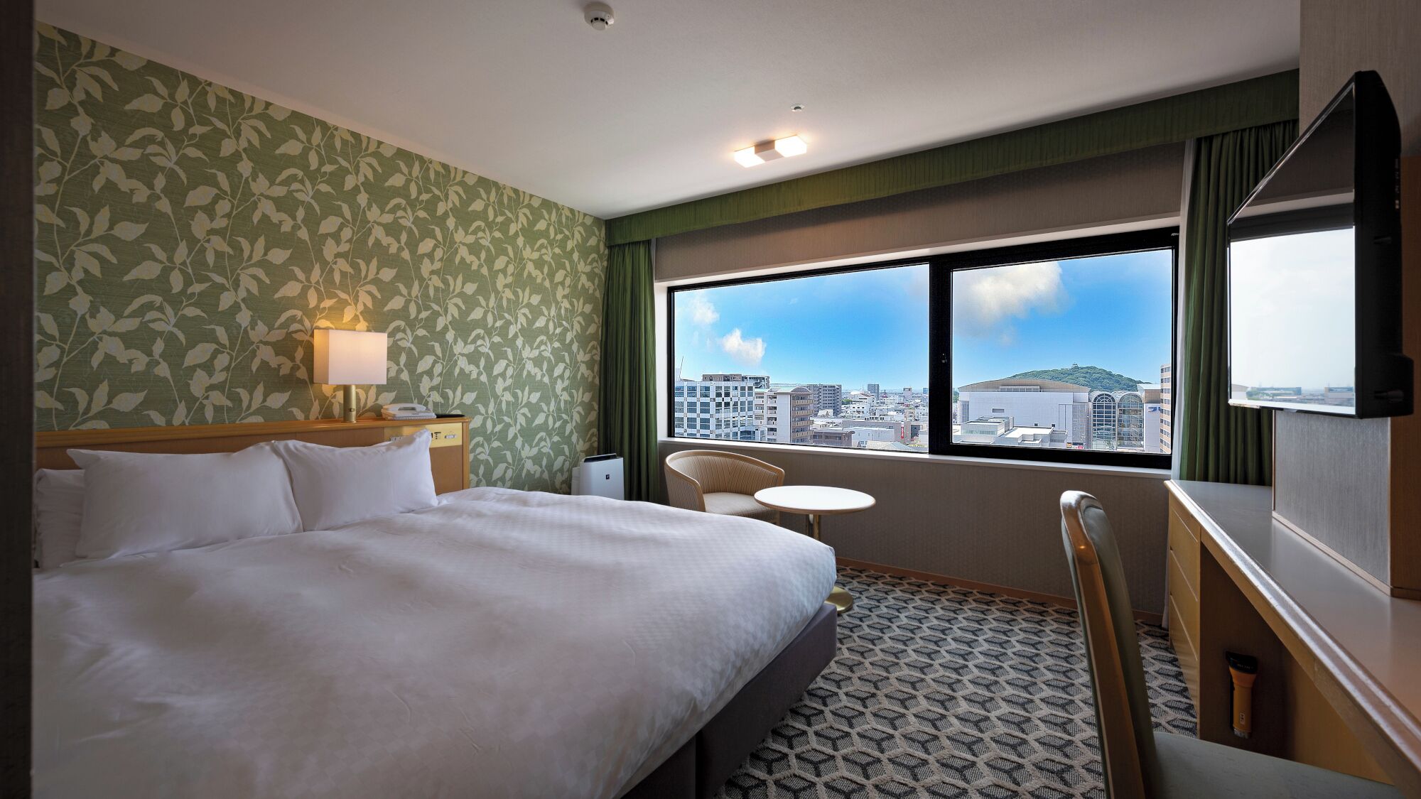 A hotel room with a bed, a chair, a small table, a lamp, a window with a city view, and a ceiling light.