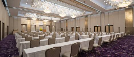 A large conference room with rows of tables and chairs, high ceilings with geometric chandeliers, and a patterned carpet.