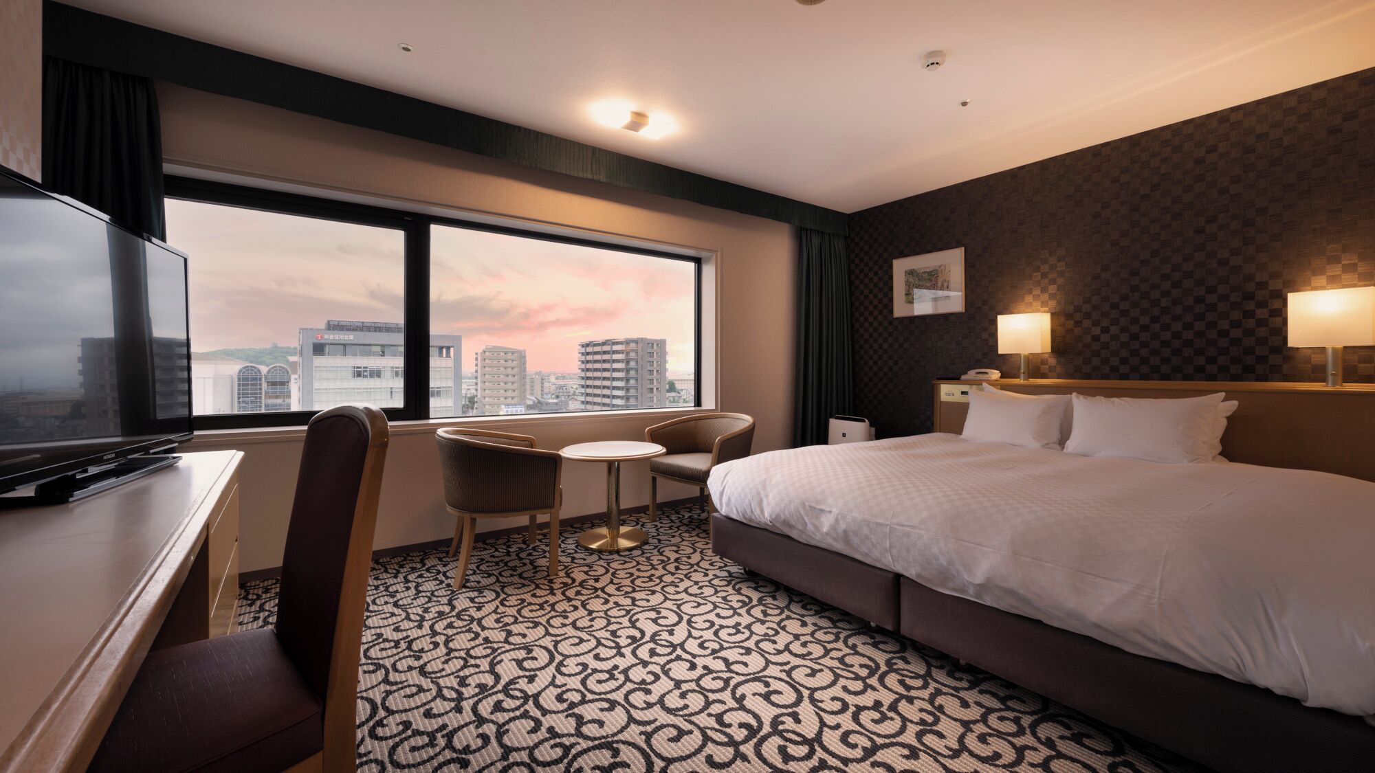 A hotel room with a large bed, a desk with a TV, a chair, a small table, and a view of the cityscape through large windows.